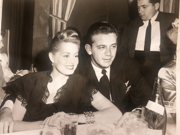 Black and white photograph of a woman with styled hair and a dark dress sitting next to a man in a suit at a table, with drinks and other people in the background.